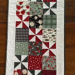 K&ouml;nnte beinhalten: Ein wei&szlig;er Tischl&auml;ufer mit grauer Umrandung zeigt ein Patchwork-Design aus roten, gr&uuml;nen, schwarzen und wei&szlig;en Stoffquadraten mit verschiedenen festlichen Mustern. Der L&auml;ufer besteht aus einem sich wiederholenden Muster von Windm&uuml;hlenformen.