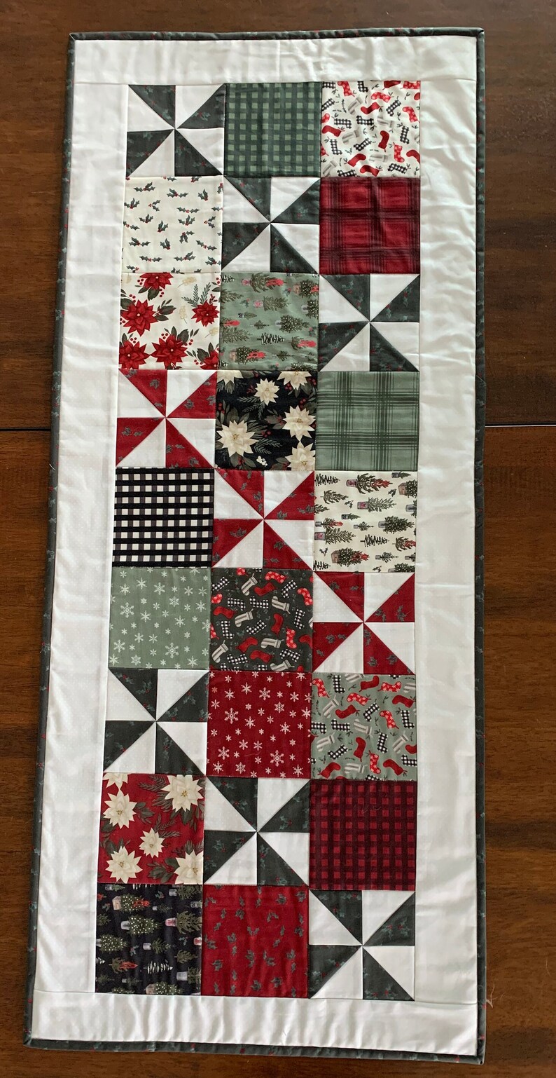 K&ouml;nnte beinhalten: Ein wei&szlig;er Tischl&auml;ufer mit grauer Umrandung zeigt ein Patchwork-Design aus roten, gr&uuml;nen, schwarzen und wei&szlig;en Stoffquadraten mit verschiedenen festlichen Mustern. Der L&auml;ufer besteht aus einem sich wiederholenden Muster von Windm&uuml;hlenformen.