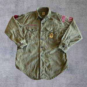 May include: Olive green button-down shirt with two chest pockets and patches. Patches include the text "FULLER CALIF" and the number "93". The shirt has a collar and buttoned cuffs. A vintage style garment.