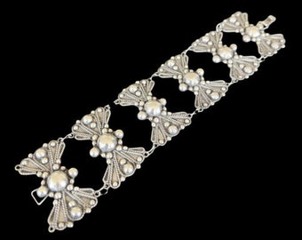 Puede incluir: Un brazalete de plata con un patrón repetitivo de formas de flores estilizadas. Cada flor tiene un centro redondo y está rodeada de círculos más pequeños en relieve.