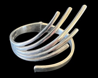 May include: A silver-toned cuff bracelet with a unique, open design. The bracelet features multiple curved bands that wrap around the wrist, with vertical bars extending upwards. The metal has a polished finish, reflecting light against a black background.