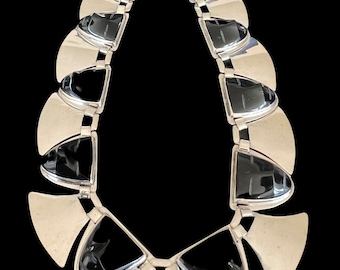 May include: A silver-toned necklace with a geometric design. The necklace features alternating silver and black elements, with a teardrop shape. The necklace is displayed against a black background, highlighting its modern aesthetic.