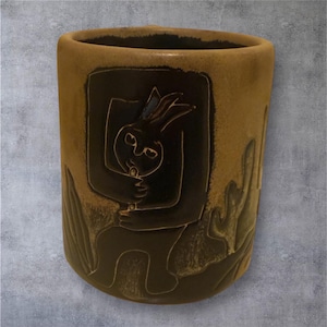 May include: A cylindrical ceramic vase in shades of brown and tan. A dark brown rectangular panel features a stylized illustration of a figure embracing a rabbit. The vase has a textured surface and a slightly flared top, with carved plant-like designs.