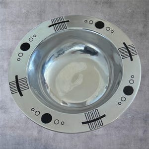 May include: A silver-colored bowl with a wide rim, decorated with black geometric patterns. The rim features a series of black circles and rectangular shapes. The bowl's interior reflects the surrounding environment, creating a shiny appearance.
