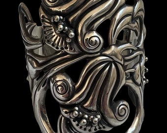 May include: A wide, silver-toned cuff bracelet with an intricate, flowing design. The openwork metalwork features swirling patterns, floral elements, and small, round accents. The bracelet has a polished finish and a bold, artistic aesthetic.