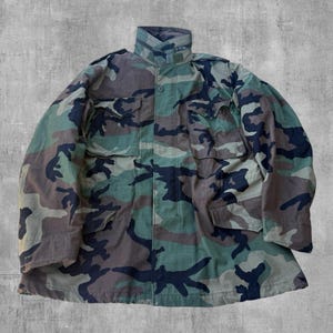 May include: A camouflage jacket in shades of green, brown, and navy blue. The jacket has a high collar, multiple pockets, and a button-up front. The military-style jacket is made of a durable fabric.