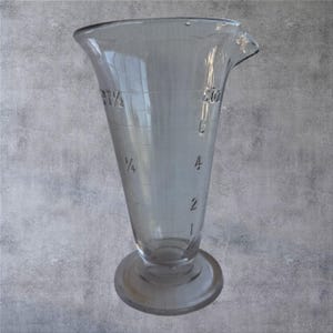 May include: A clear glass measuring beaker with a flared top and a round base. The beaker has measurement markings in ounces and fractions, with numbers 1, 2, 4, and 8 oz. The glass has a spout for easy pouring.