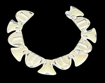 May include: A silver-toned necklace with a geometric design. The necklace features multiple connected, fan-shaped elements with a polished finish. The design is symmetrical and rests against a black background, highlighting the jewelry's reflective surface.