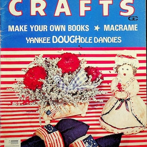 May include: The cover of Creative Crafts magazine, August 1976, features a patriotic theme. It includes a floral arrangement with red flowers, a doll, and napkin rings decorated with American flags. The magazine's title is in large, white letters.