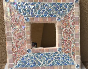 May include: A square mirror with a mosaic frame made of small, colorful glass tiles. The frame features a blue and pink design with a wavy pattern. The mirror is surrounded by a border of small, white glass tiles.