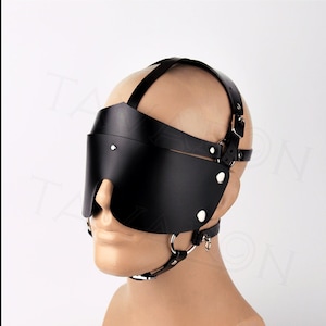 May include: Black leather blindfold with a head strap and a nose ring. The blindfold is worn on a mannequin head.