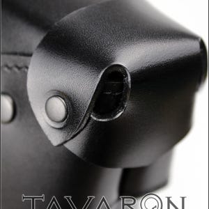 Puede incluir: Primer plano de un producto de cuero negro con una superficie texturizada. La imagen destaca las costuras, los remaches y una solapa curva asegurada con un broche. La palabra "TAVARON" est&aacute; impresa en una fuente serif en la parte inferior.