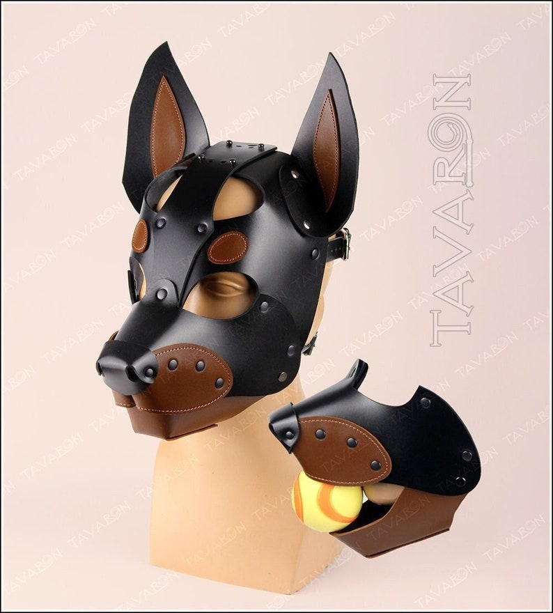 May include: Black leather dog mask with brown leather accents and metal studs. The mask has a snout with a brown leather nose and a small opening for the mouth. The mask also has two brown leather ears.