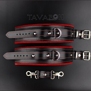 May include: Black leather restraints with red velvet lining and silver hardware. The restraints have a D-ring and a buckle closure. The restraints are connected by a short chain with two silver clips.