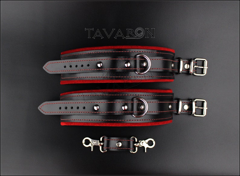 May include: Black leather restraints with red velvet lining and silver hardware. The restraints have a D-ring and a buckle closure. The restraints are connected by a short chain with two silver clips.
