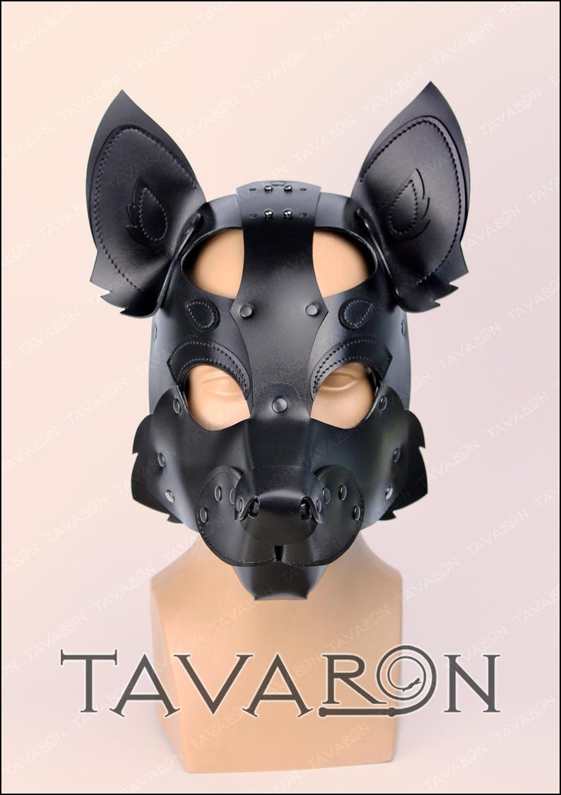 Puede incluir: Una m&aacute;scara de cuero negro estilo perro con orejas grandes y erguidas y un hocico. La m&aacute;scara presenta detalles de remaches y est&aacute; dise&ntilde;ada para cubrir la cara. La marca "TAVARON" se muestra en la parte inferior.