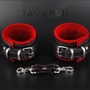 May include: A pair of black leather cuffs with red velvet lining and silver buckles. The cuffs are connected by a black leather strap with a silver hook and eye closure. The text "TAVARON" is visible in the background.