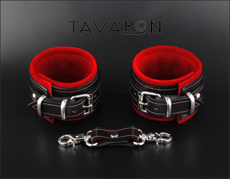 May include: A pair of black leather cuffs with red velvet lining and silver buckles. The cuffs are connected by a black leather strap with a silver hook and eye closure. The text "TAVARON" is visible in the background.