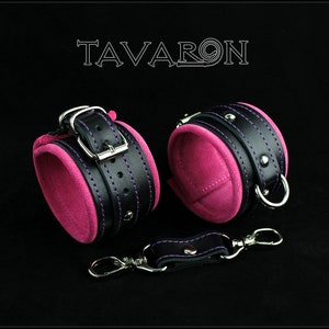 May include: A pair of black leather cuffs with pink velvet lining and purple stitching. The cuffs have silver buckles and metal rings. A black leather strap with a silver hook is included.