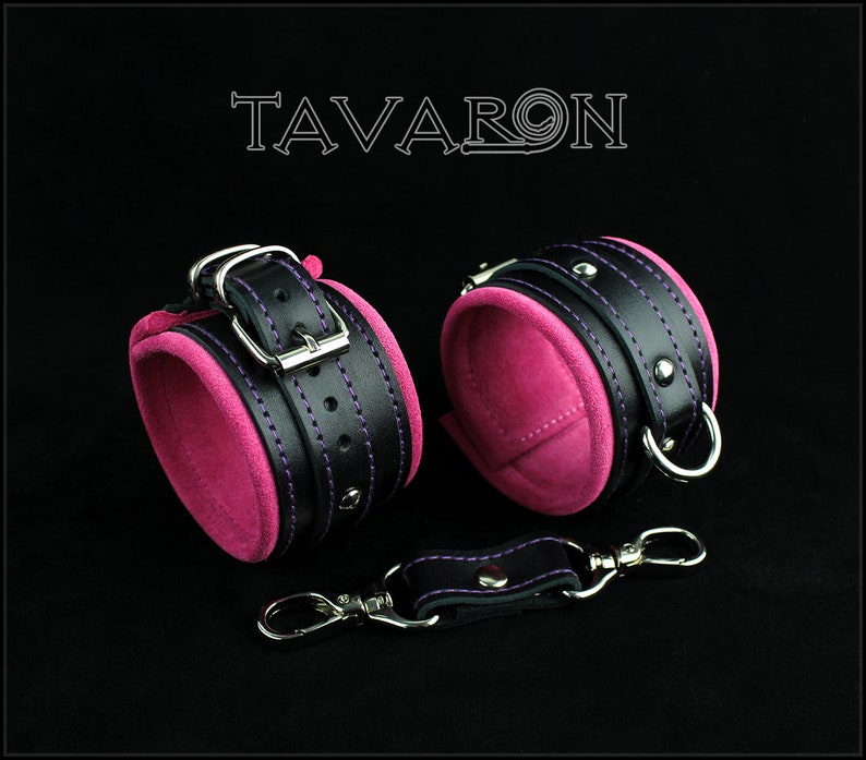 May include: A pair of black leather cuffs with pink velvet lining and purple stitching. The cuffs have silver buckles and metal rings. A black leather strap with a silver hook is included.