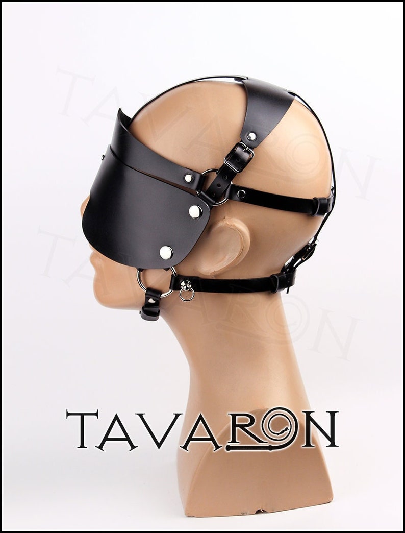 May include: Black leather blindfold with a harness that goes around the head. The blindfold has a strap that goes under the chin and a ring on the strap. The harness has a strap that goes over the top of the head and two straps that go around the back of the head.
