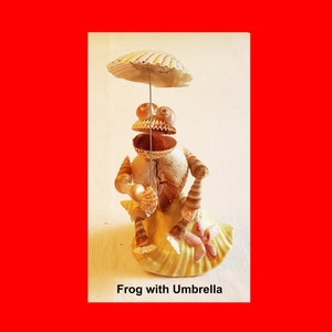 May include: A whimsical frog sculpture crafted from seashells, holding a seashell umbrella. The frog is seated on a large shell base, with the text "Frog with Umbrella" below the image. The sculpture is set against a red background.