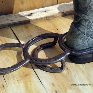 May include: A metal boot jack made from two horseshoes welded together. The boot jack is sitting on a wooden surface. The boot jack is a rustic and unique way to remove boots.