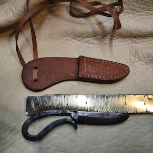 May include: A hand-forged knife with a curved handle and a leather sheath. The knife is approximately 6 inches long, with a blade that is about 2 inches long. The sheath is brown and has a stitched design. A leather strap is attached to the sheath.