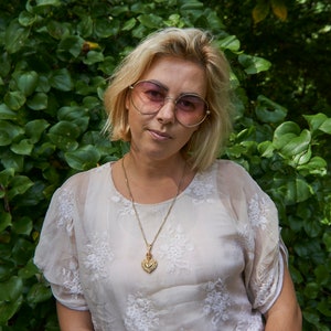 May include: A woman with blonde hair wearing heart-shaped sunglasses and a white blouse with delicate embroidery. She is wearing a gold chain necklace with a heart-shaped pendant.