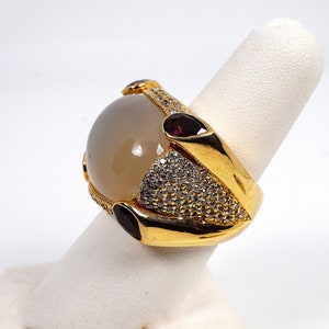 May include: A gold ring with a large, round, white gemstone surrounded by smaller, clear gemstones. The ring also has three small, red gemstones on the side.