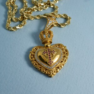 May include: A gold heart-shaped pendant with a crown and a cross set with red and white stones hangs from a gold rope chain.