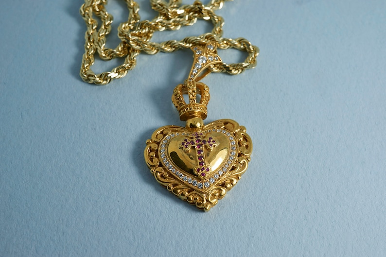May include: A gold heart-shaped pendant with a crown and a cross set with red and white stones hangs from a gold rope chain.