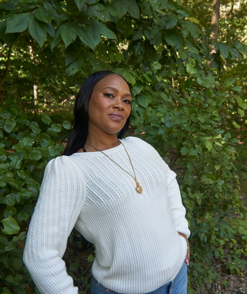 May include: A woman wearing a white knitted sweater with a gold chain necklace and a heart-shaped pendant. She is standing in front of a green leafy background.