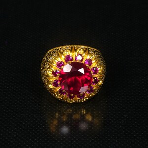 Könnte beinhalten: Ein goldener Ring mit einem großen, runden, roten Edelstein, der von kleineren roten Edelsteinen umgeben ist. Der Ring hat ein detailliertes, verziertes Design.