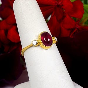 Può includere: Un anello d'oro con una grande pietra di rubino ovale e due pietre bianche rotonde più piccole. L'anello è su un espositore bianco.