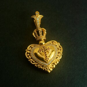 May include: A gold heart-shaped pendant with a crown on top and a cross in the center. The pendant is decorated with small white and red gemstones.