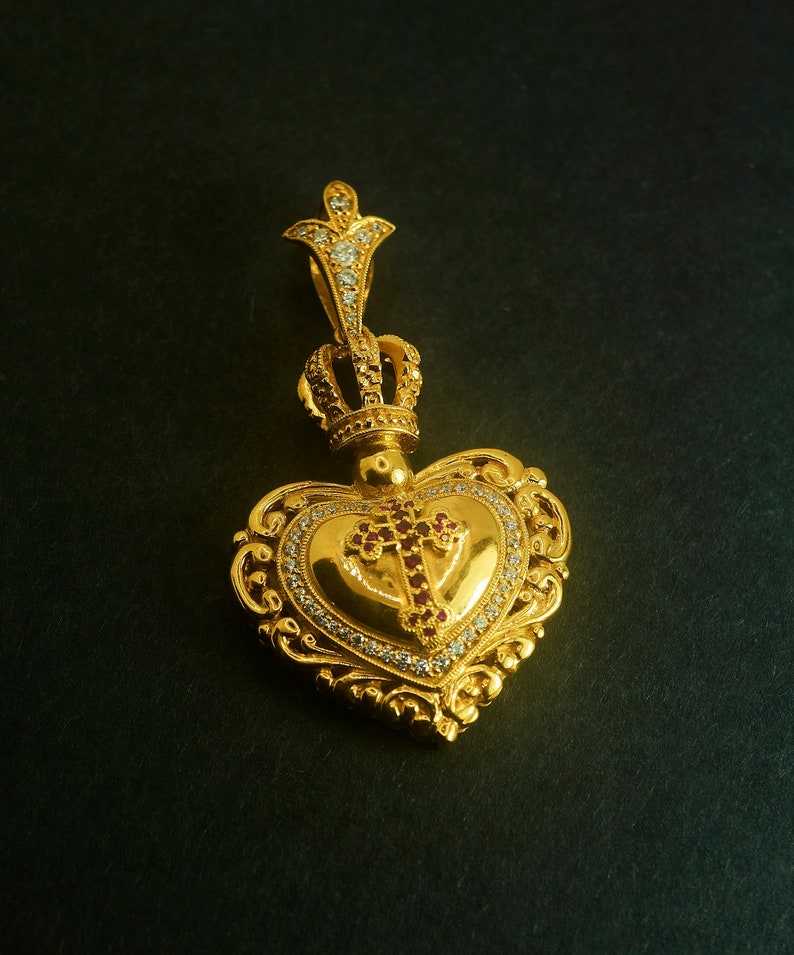 May include: A gold heart-shaped pendant with a crown on top and a cross in the center. The pendant is decorated with small white and red gemstones.