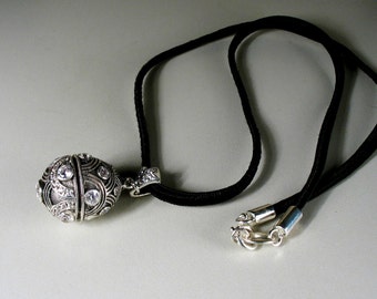 May include: A silver pendant necklace with a round, ornate charm featuring clear rhinestones. The necklace is on a black cord with a silver clasp.