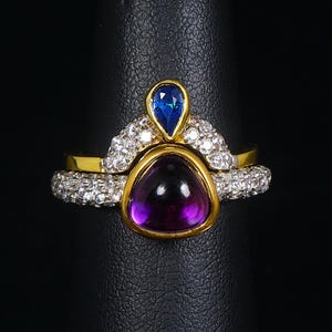 Puede incluir: Un anillo de oro con una piedra preciosa púrpura y una piedra preciosa azul engastadas en una banda de oro con un halo de diamantes.