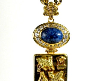 May include: Gold-toned necklace with a large oval blue stone pendant surrounded by clear crystals. The pendant is connected to a square charm with embossed winged creatures and architectural elements. The chain is a thick, textured gold.
