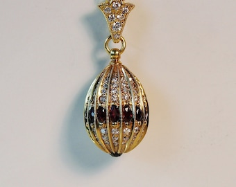 May include: A gold pendant necklace with a round, egg-shaped charm. The charm is decorated with small, clear gemstones and five red gemstones.