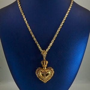 May include: A gold rope chain necklace with a heart-shaped pendant. The pendant features a crown, a cross, and intricate detailing. The necklace is displayed on a blue velvet stand.