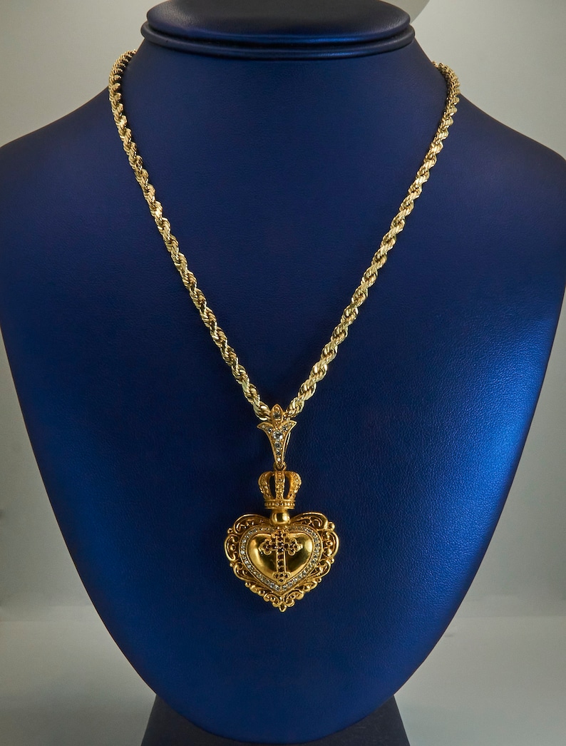 May include: A gold rope chain necklace with a heart-shaped pendant. The pendant features a crown, a cross, and intricate detailing. The necklace is displayed on a blue velvet stand.