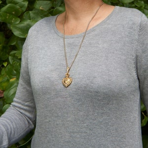 May include: A silver chain necklace with a gold heart pendant. The pendant has a detailed design and is worn by a person wearing a gray sweater.