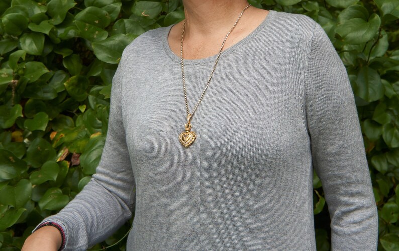May include: A silver chain necklace with a gold heart pendant. The pendant has a detailed design and is worn by a person wearing a gray sweater.
