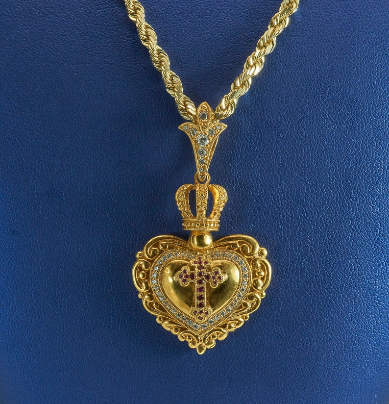 May include: A gold heart-shaped pendant with a crown and a cross set with red and white stones hangs from a gold rope chain.