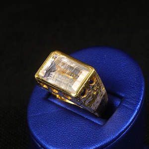 May include: A gold ring with a large, rectangular, clear gemstone. The ring has a textured band with decorative cutouts. The gemstone is set in a gold bezel. The ring is displayed on a blue surface against a black background.