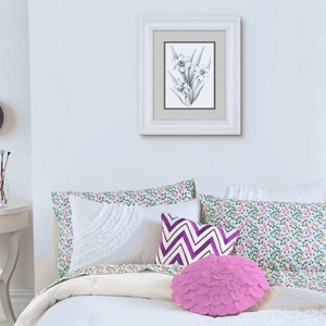May include: A white bed with a white, green, and purple floral patterned duvet cover and pillowcases. There are three pillows on the bed, one with a white ruffle, one with a purple and white chevron pattern, and one pink ruffled pillow. A framed black and white drawing of a flower hangs on the wall above the bed.
