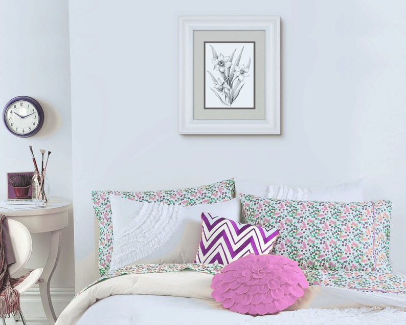 May include: A white bed with a white, green, and purple floral patterned duvet cover and pillowcases. There are three pillows on the bed, one with a white ruffle, one with a purple and white chevron pattern, and one pink ruffled pillow. A framed black and white drawing of a flower hangs on the wall above the bed.