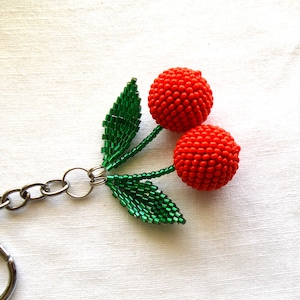 May include: A beaded cherry keychain with a silver chain and green leaves. The cherries are red and made of small beads.